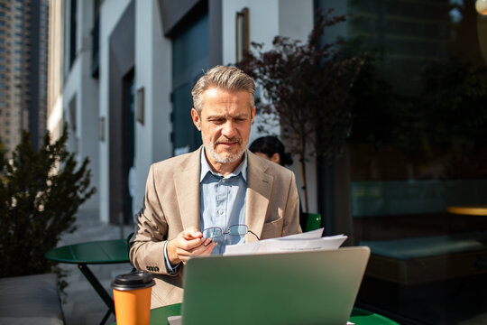 Mature businessman reviewing documents on laptop at outdoor cafe