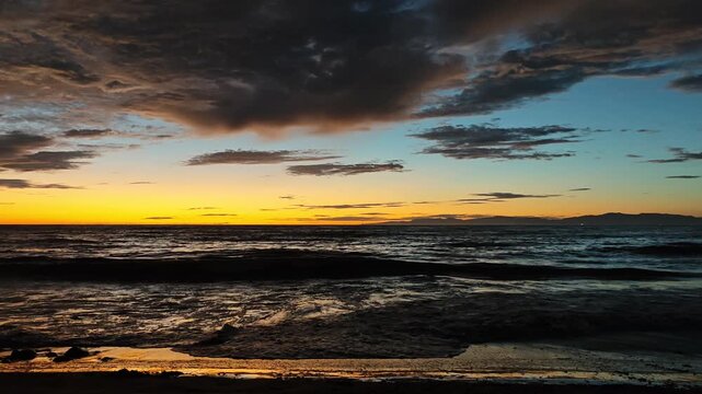 The sun dips below the horizon casting a warm glow on the sea as waves gently roll onto a pebble beach