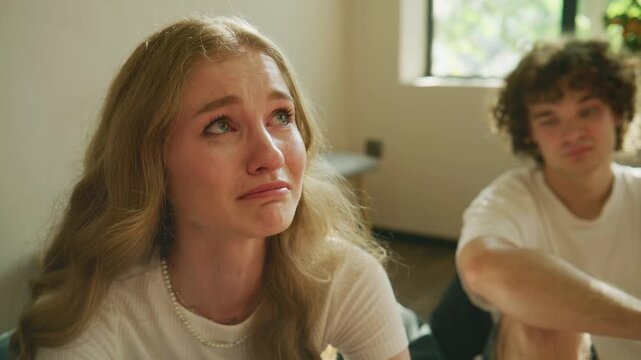 A young woman sits in a therapy room, showing visible emotions while crying. Another person is present, observing her reactions during the session.
