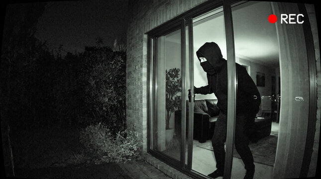 Masked burglar in black hoodie entering house through sliding glass door at night. Home break-in caught on surveillance camera. Theft, robbery and home security concept.