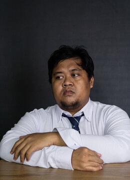 A young Asian male wearing a white long sleeved shirt and blue necktie sits at a wooden table against a dark studio background. The corporate worker rests crossed arms on the desk