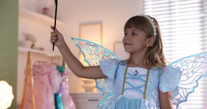 Smiling little girl in fairy costume with wings and headband playing with magic wand at home. Camera moving forward and backwards