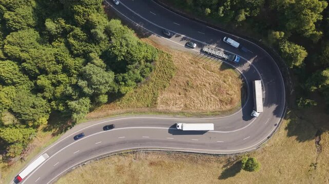 Top-down aerial view of trucks and cars navigating a sharp hairpin bend on a winding mountain road surrounded by dense green forest on a sunny summer day.