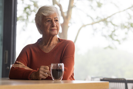 Senior woman sitting at wood table by window wearing rust sweater, holding glass mug, gazing right