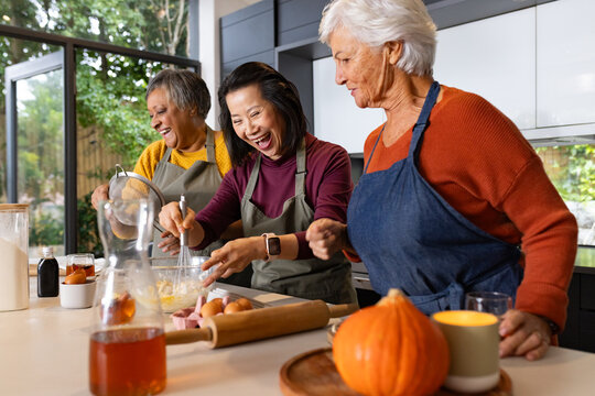 Diverse female friends wearing aprons whisking batter at kitchen island, using glass bowl, whisk