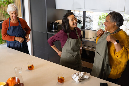 Diverse senior female friends laughing in aprons making iced drinks near pumpkin on kitchen island