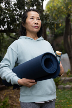 Senior Asian woman standing in garden wearing light blue hoodie and holding yoga mat, water bottle