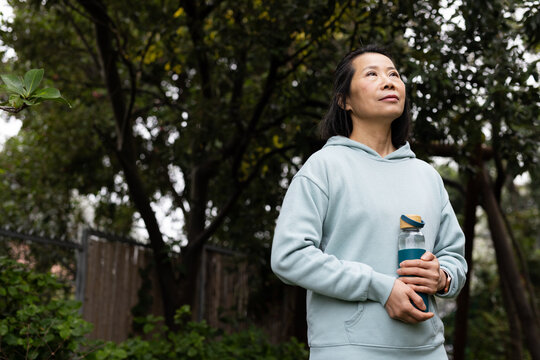 Senior Asian woman standing in backyard holding reusable bottle wearing blue hoodie, copy space