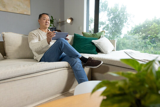 Senior Asian man sitting on beige sectional sofa at home holding tablet and looking at screen