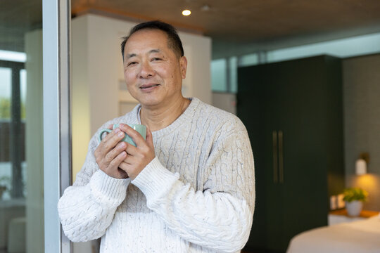 Senior Asian man standing in bedroom holding blue-green mug wearing cream knit sweater