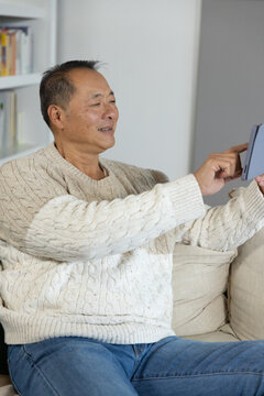Senior Asian man wearing cable-knit sweater and jeans holding tablet, tapping screen in living room