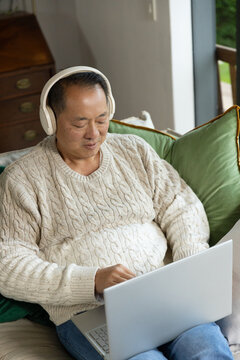 Senior Asian man sitting on sofa wearing cream cable-knit sweater and headphones using laptop