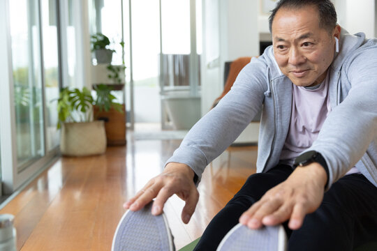 Senior Asian man stretching reaching shoes on exercise mat wearing watch in living room, copy space
