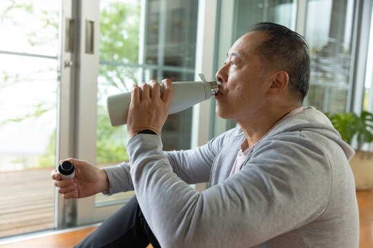 Senior Asian man sitting on wooden floor by glass doors sipping insulated bottle, wearing hoodie