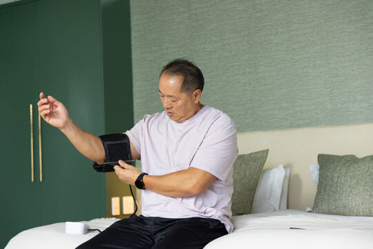 Senior Chinese man sitting on bed adjusting black blood pressure cuff, watching white monitor