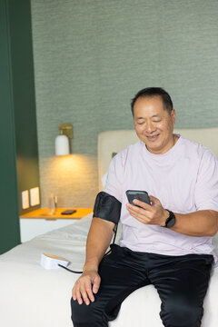 Senior Chinese man sitting on bed in room, wearing tee and checking cuff monitor using smartphone