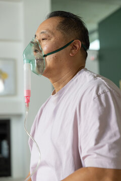 Senior Asian man wearing oxygen mask with green strap and pink tubing connector at home