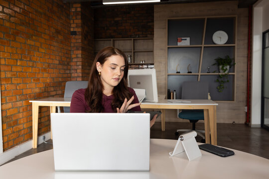 Adult woman wearing maroon top holding tablet, working on silver laptop with smartphone in office