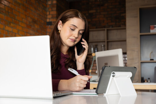 Female worker sitting at desk wearing burgundy top and gold hoops, using phone and writing notes