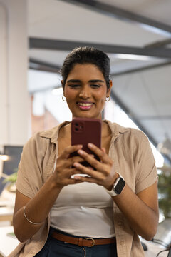 Indian woman standing in office holding maroon phone, wearing smartwatch and hoop earrings