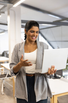 Indian woman holding silver laptop, gesturing in open-plan office with desks, wearing gray blazer