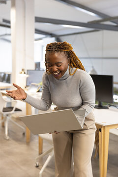 Mid-30s woman standing in open office holding silver laptop, wearing grey sweater, gesturing