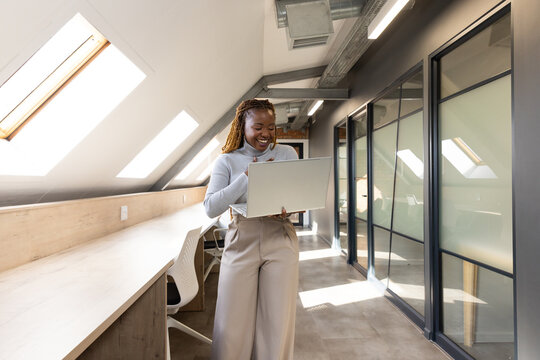African woman standing holding laptop in loft with skylights, glass offices in turtleneck