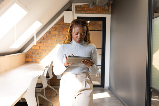 African woman standing using tablet in loft office under skylights wearing gray top and beige pants