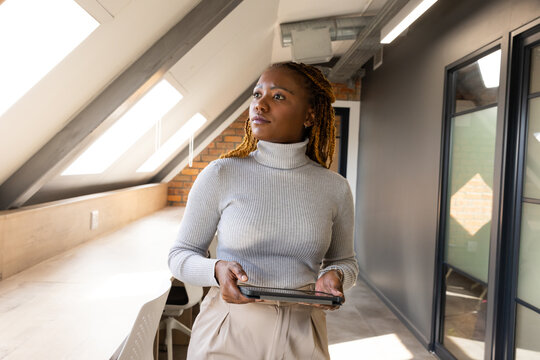 African American mid adult female standing and holding tablet, looking right in loft corridor