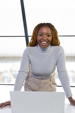 African woman leaning over laptop at modern office desk, smiling in gray turtleneck and bracelets