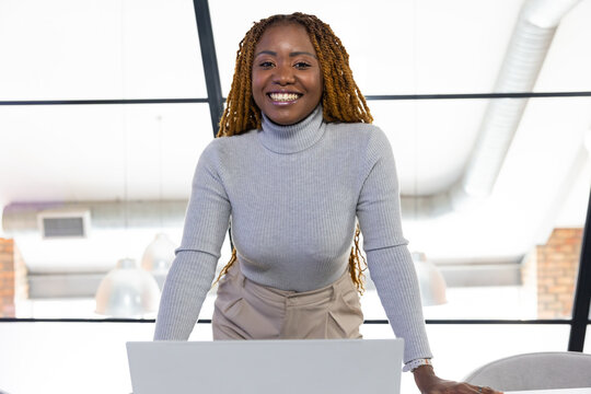 Woman leaning over desk, smiling while using laptop in loft office, wearing gray turtleneck