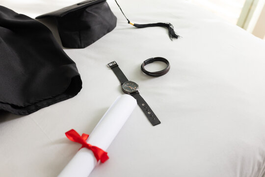 Diploma tied with red ribbon is resting on white bedsheet, cap, gown fabric, watch and bracelet