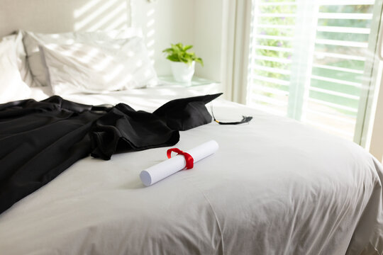 rolled diploma tied red ribbon lying on sheets, gown cap draping shutters casting shadows, plant