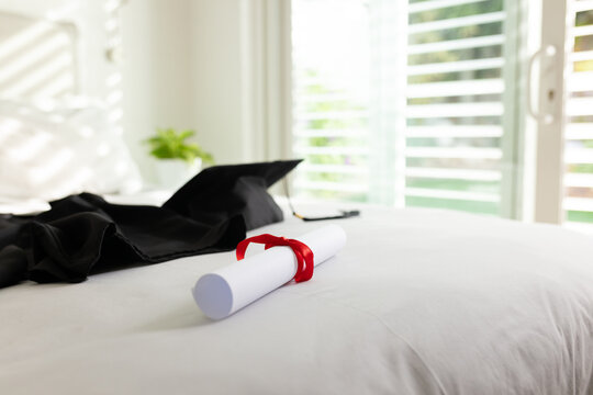Rolled diploma scroll resting on white bed linens near black gown and cap, sunlight filling room
