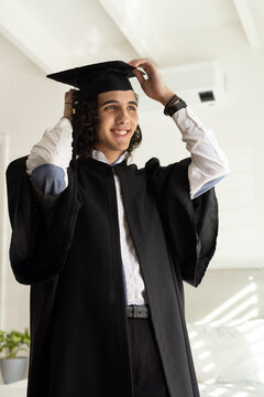 Man in 20s adjusting mortarboard cap and smiling, wearing graduation gown in bedroom