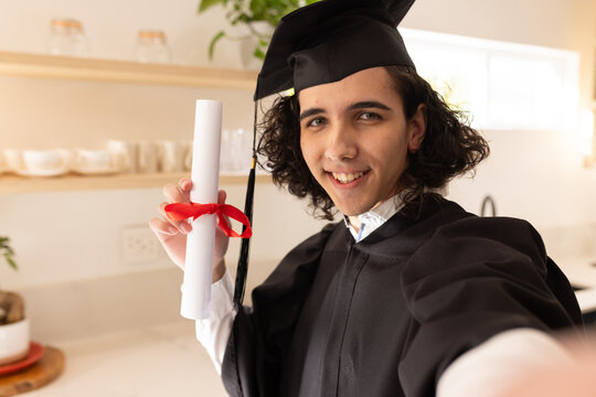 African American male holding diploma taking selfie with phone in home kitchen wearing gown and cap