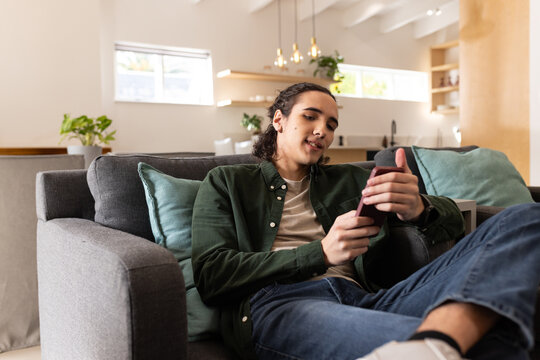 Man reclining on gray upholstered sofa, checking smartphone in modern living room with teal pillows