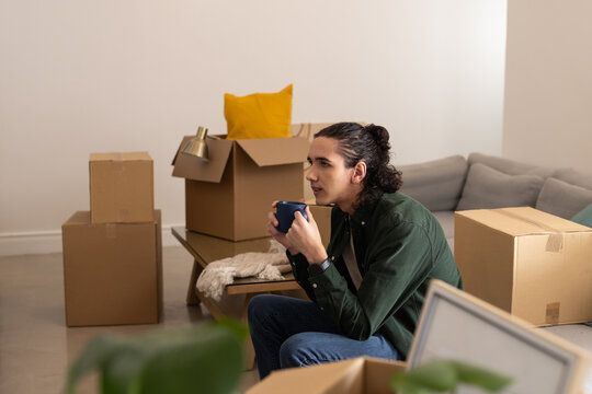 African American man sitting in living room wearing dark green shirt holding mug amid moving boxes