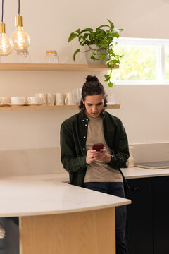 African American man in his 20s leaning on curved kitchen island, checking red smartphone