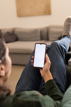 African American man reclining on sofa holding phone with blank screen at home wearing denim jeans