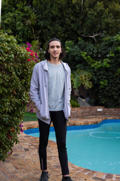 Man standing on brick deck in yard by turquoise pool, fuchsia hedge, wearing hoodie