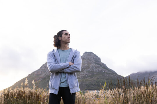African American man standing with arms crossed in dry grass field at mountain foot wearing hoodie