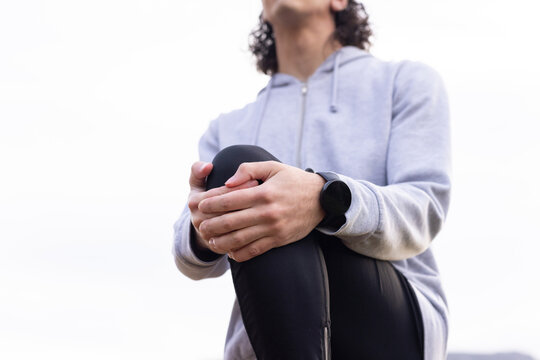 African American man sitting clasping knee against cloudy sky wearing gray hoodie leggings watch