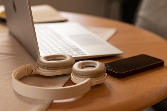 White over-ear headphones are resting on round wooden table with laptop and smartphone