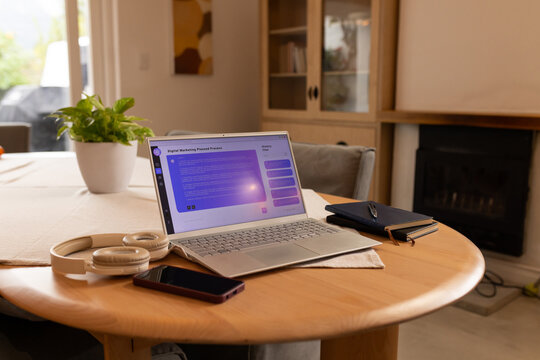 Open silver laptop is sitting on round wooden table near window with headphones and notebooks