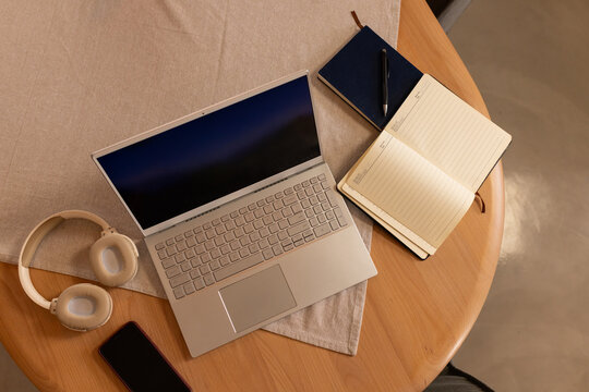 Open silver laptop with dark screen is sitting diagonally on round wooden table with notebooks