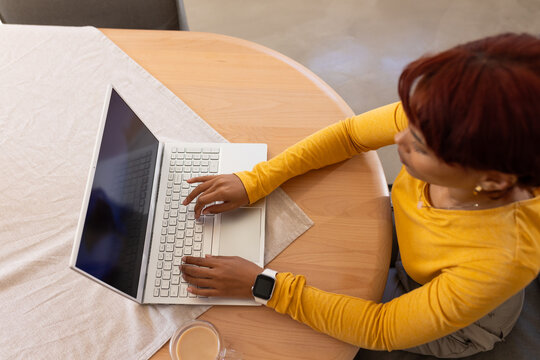African woman typing on white laptop at home on round wood table, mustard-top smartwatch