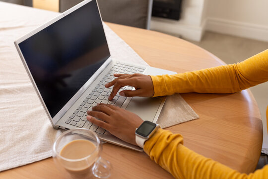 African American woman typing on silver laptop at wooden table wearing mustard top and smartwatch