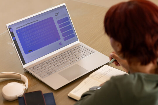 Woman sitting at wooden table dark green top writing in notebook, viewing laptop chat, copy space
