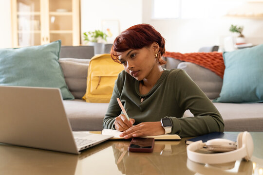 African American woman sitting in living room writing in notebook while glancing at laptop